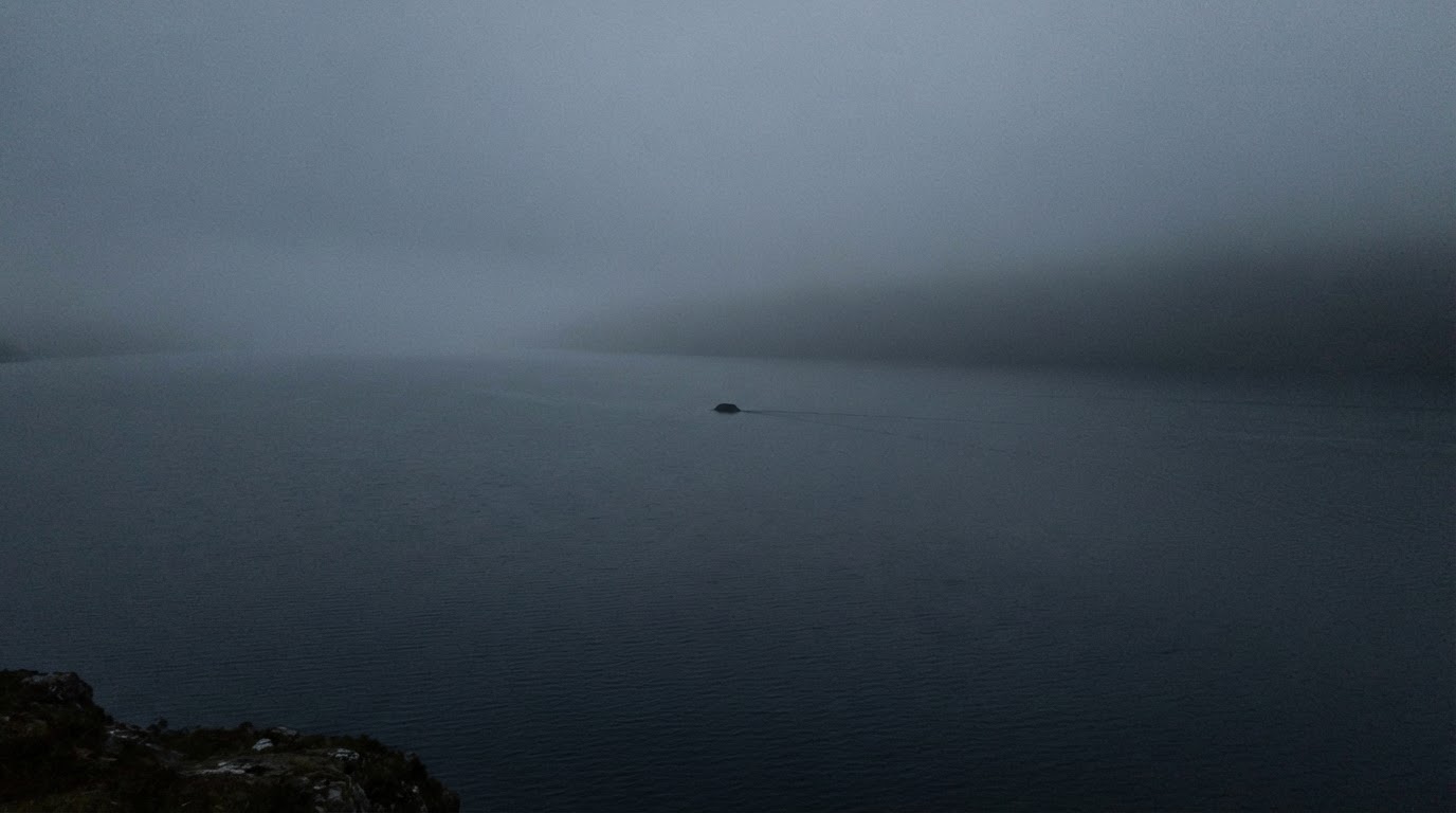 A grainy, distant telephoto-style shot of a small dark hump breaking the misty lake surface, trailing a faint wake — a recreation modeled on the sighting photos.