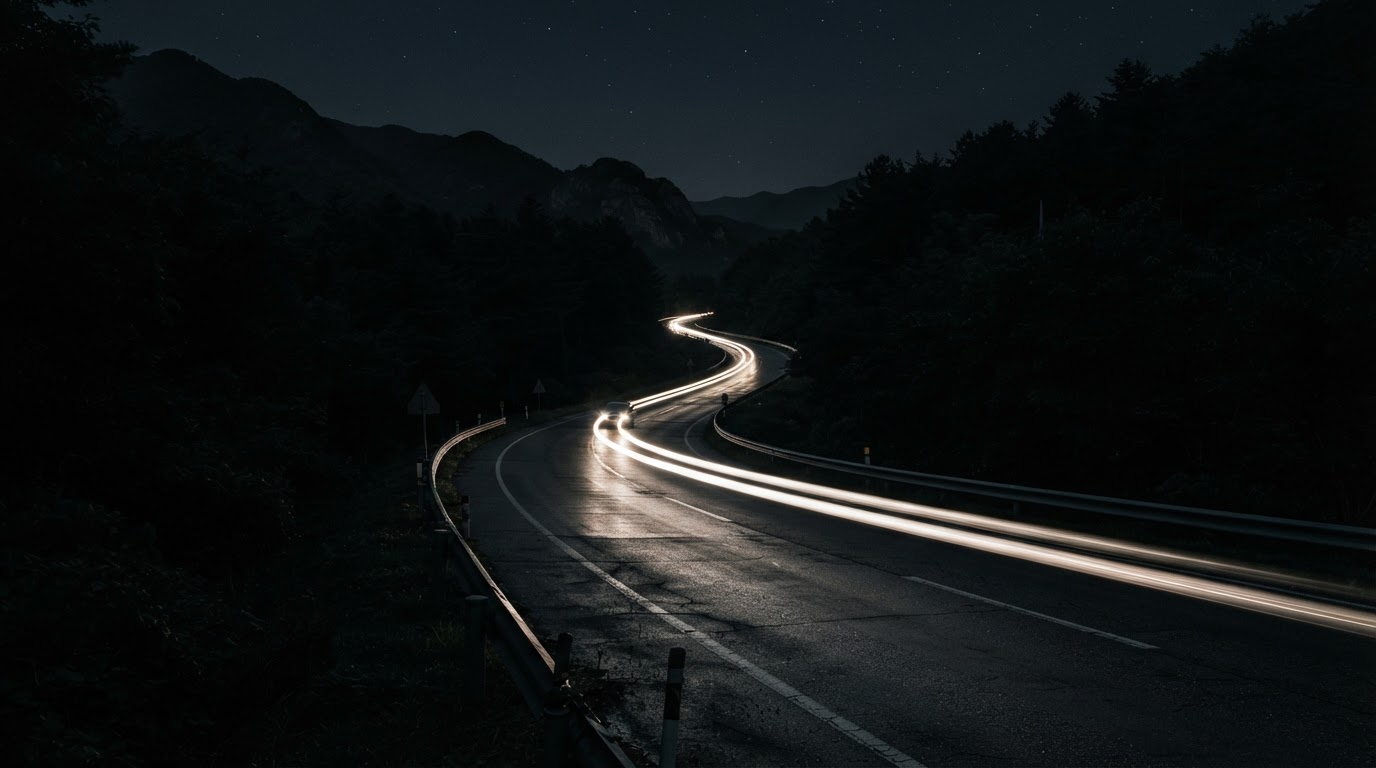 A dark, deserted provincial road with no streetlamps, only headlights smearing across the night pavement