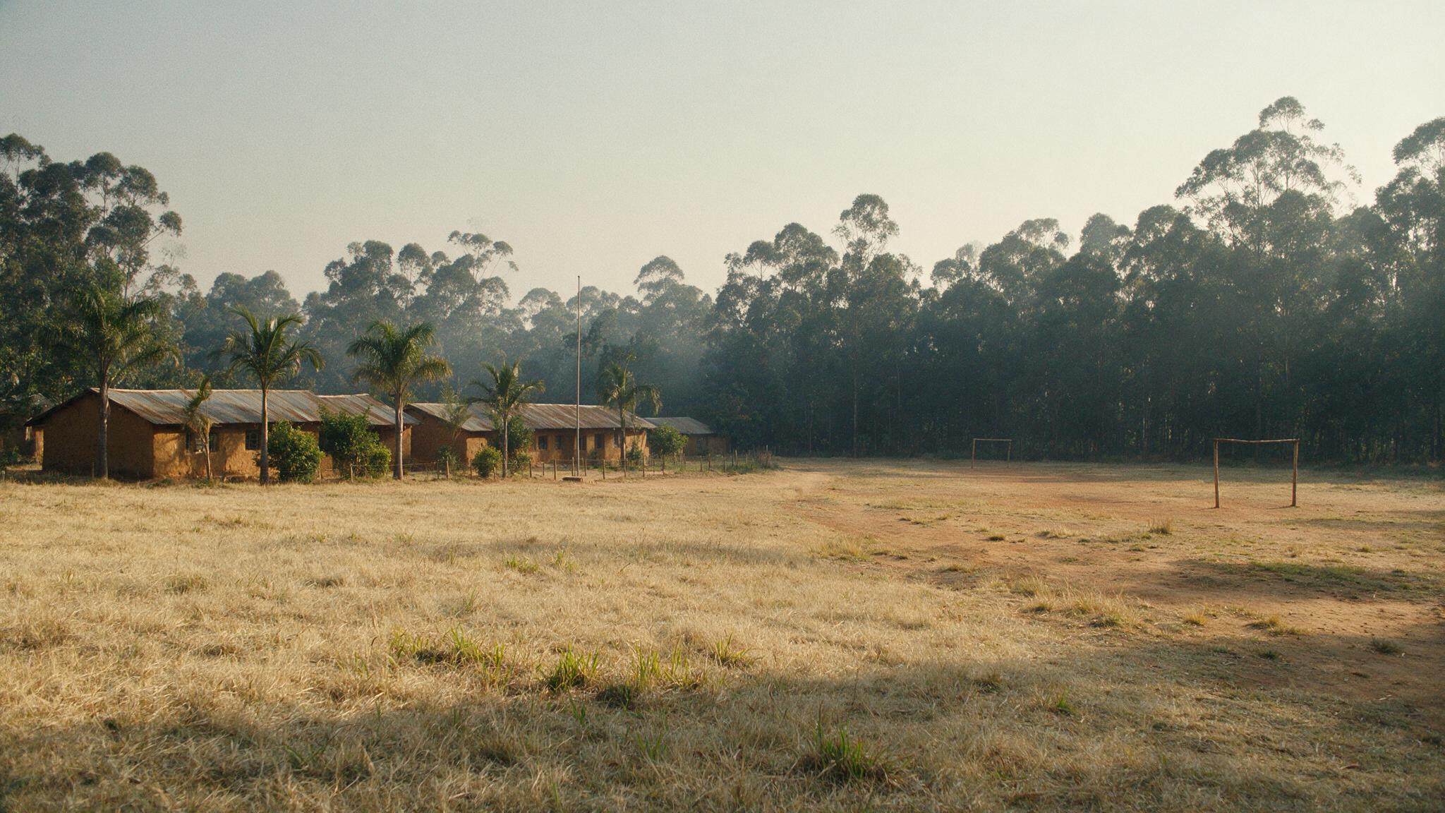 Distant view of a rural African primary school playground bordered by dry scrubland and a line of eucalyptus trees under a hazy morning sky, no people visible, photorealistic