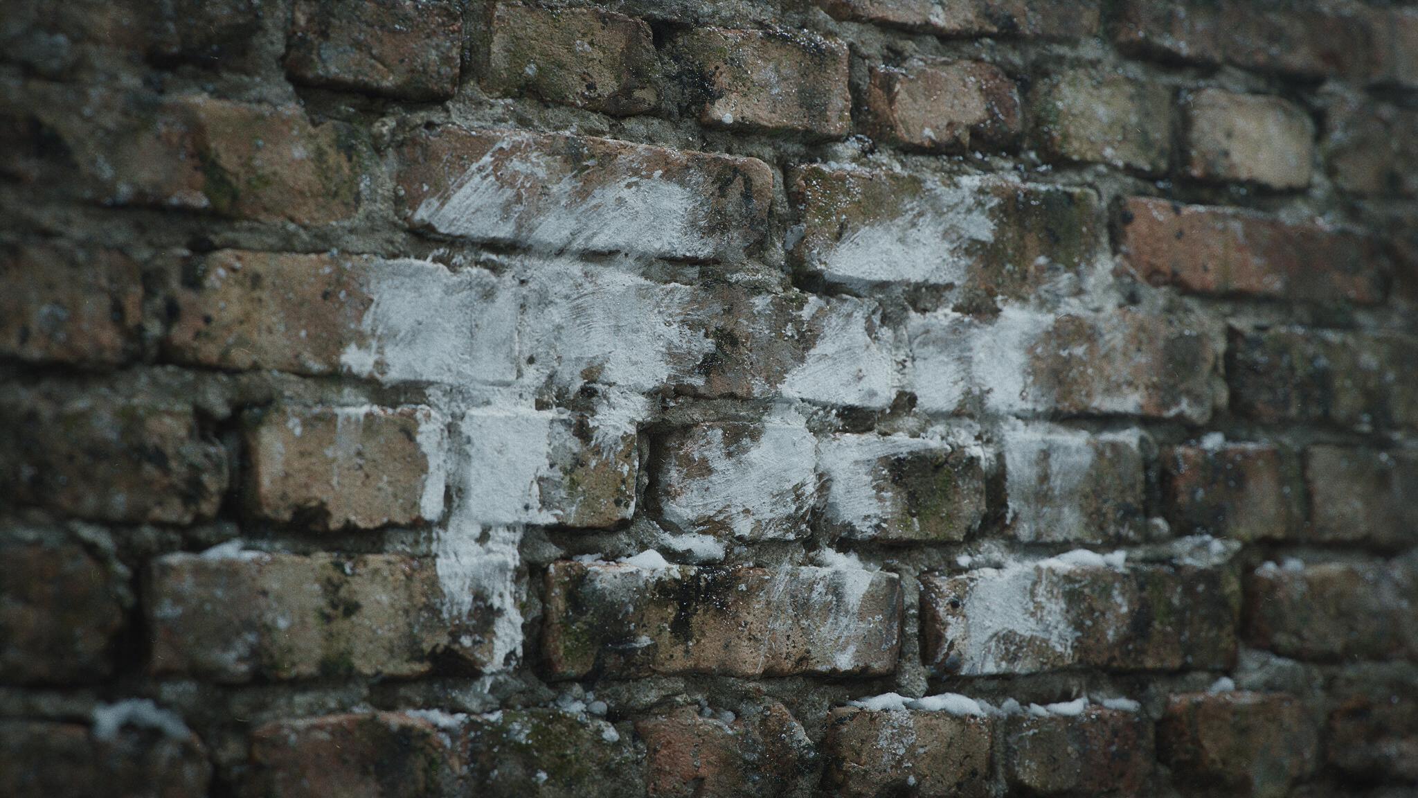 A close-up of an old, stained brick wall, faint traces of white paint smeared across its surface as though half wiped away but not readable as letters, grey winter light, an eerie, bleak and ominous mood, no people (mood recreation, not real footage)