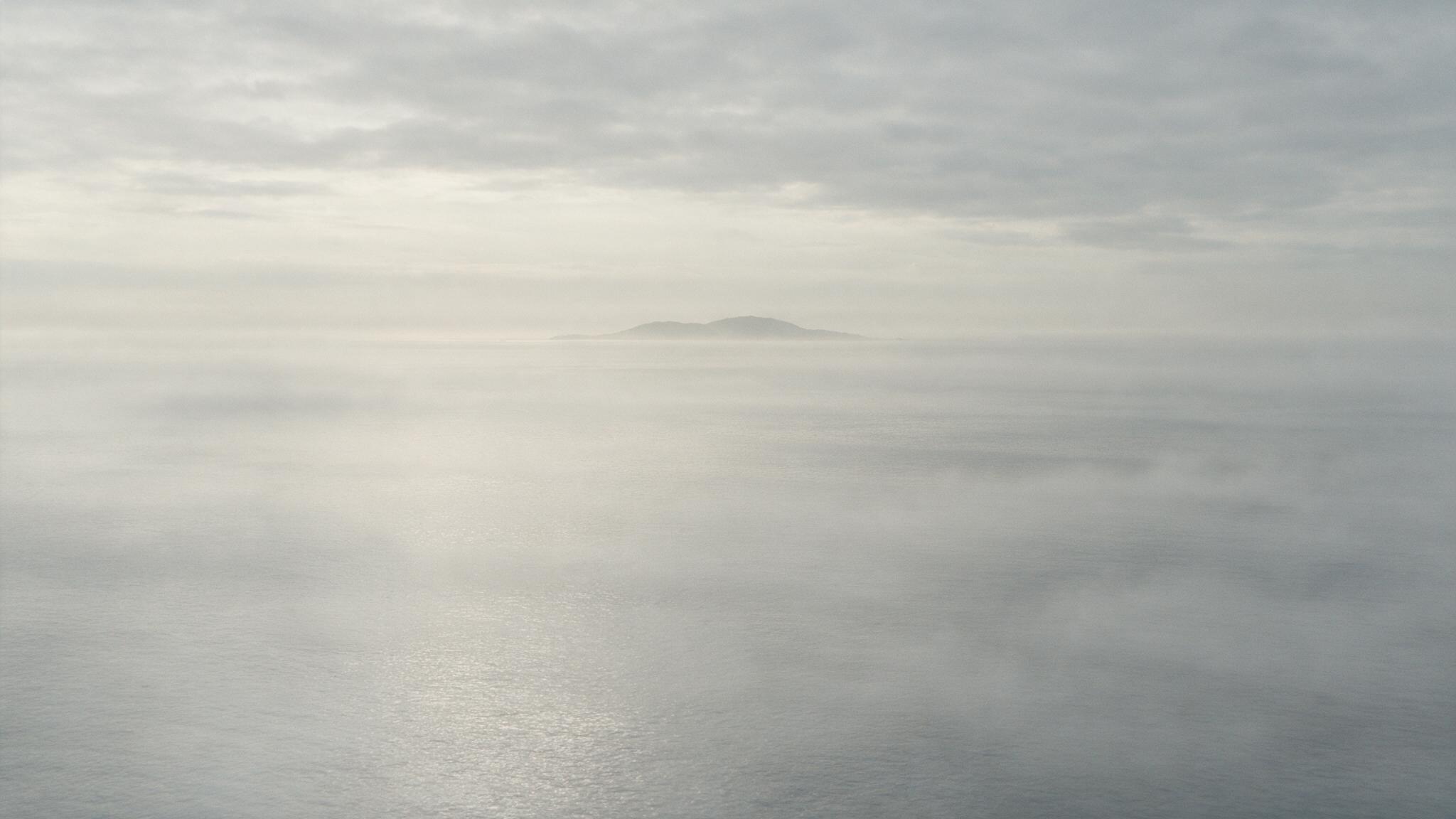A misty southern horizon, an island faintly visible in silhouette far out to sea, calm waves, a quiet and mysterious mood