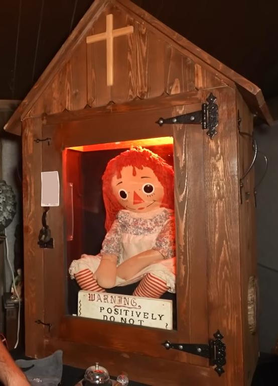 The real Annabelle doll in the Warren Occult Museum — the glass-fronted cabinet marked with a cross and the "WARNING POSITIVELY DO NOT OPEN" sign.