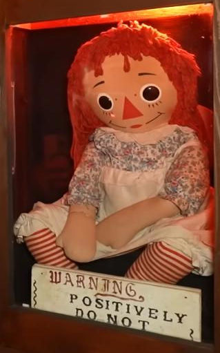 A close view of the real Annabelle inside her case — a Raggedy Ann rag doll with red yarn hair and striped stockings, the warning sign in front of her.