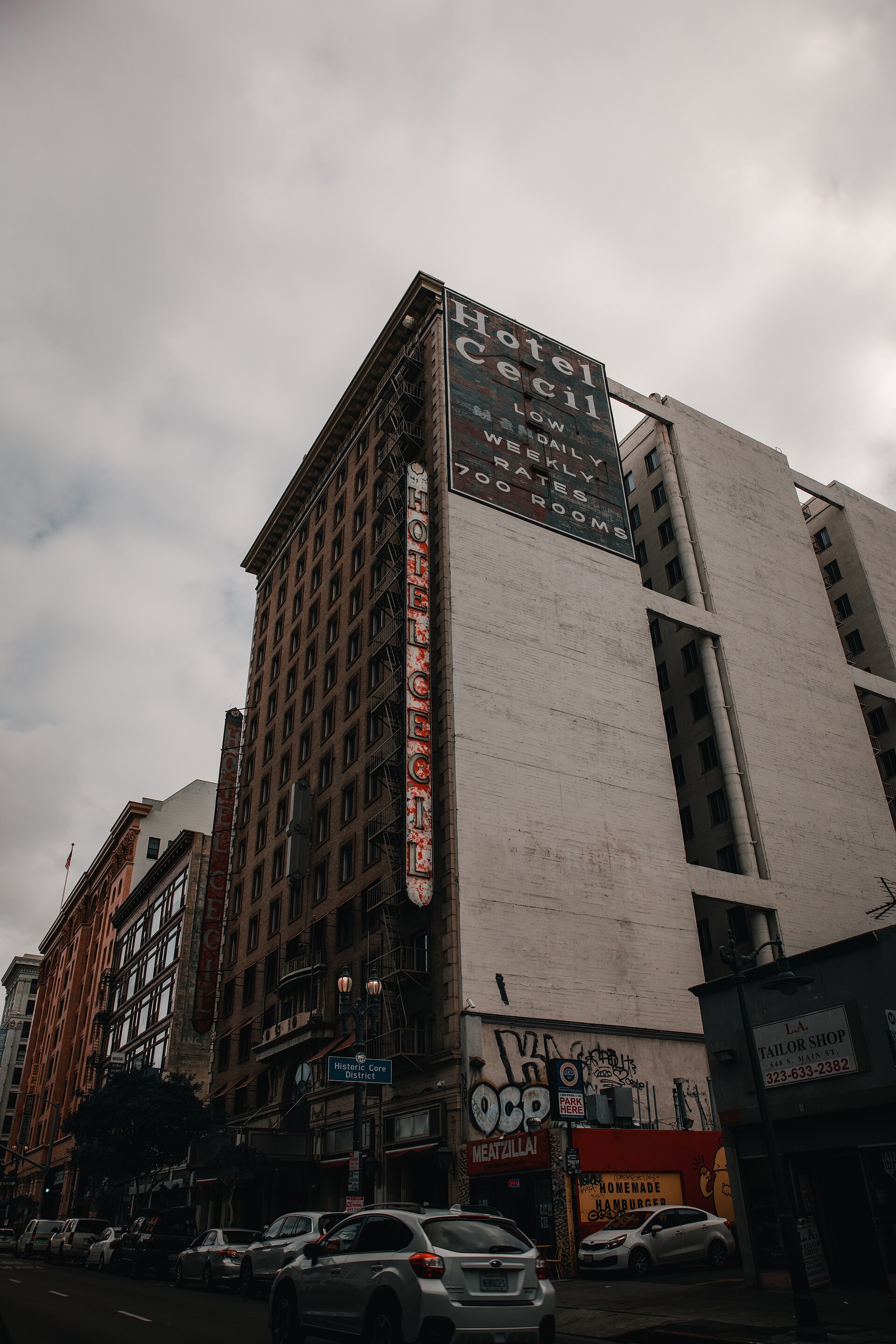 The Cecil Hotel seen from below under an overcast sky, its large rooftop sign reading "Hotel Cecil / LOW DAILY WEEKLY RATES / 700 ROOMS."