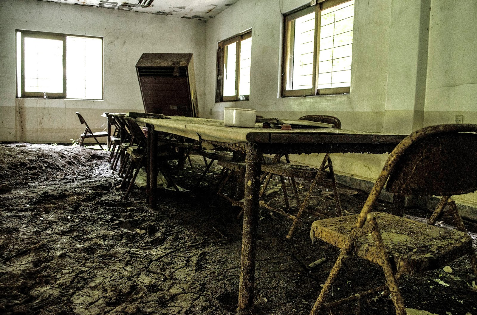 Inside the real building before demolition — a long table thick with dust, folding chairs, and a collapsed ceiling, a room left exactly as it was abandoned. Photographed on site in 2014.