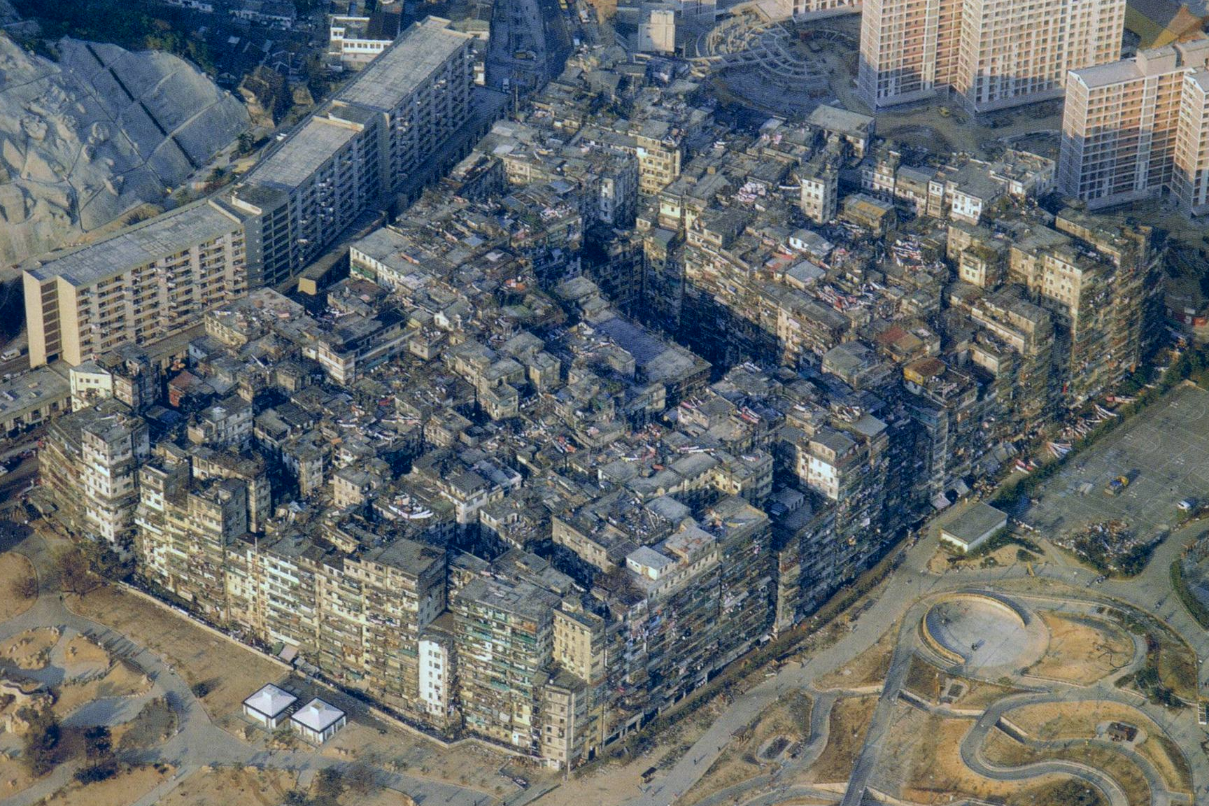 An aerial photograph of Kowloon Walled City taken in 1989, its dense block standing in sharp contrast to the modern apartment towers around it.
