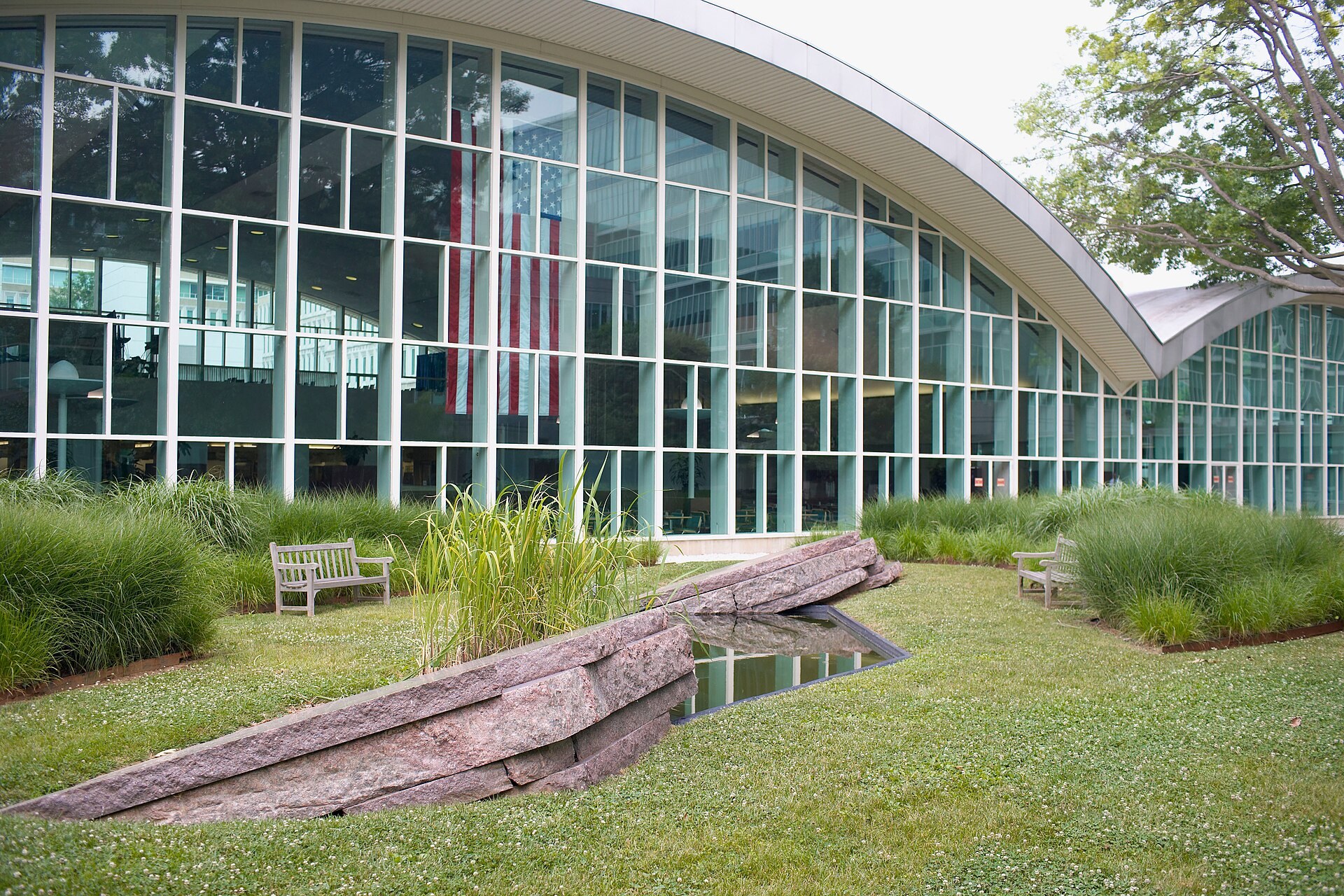 The CIA headquarters courtyard where Kryptos stands, with its split granite slabs, still reflecting pool, and the glass wall of the cafeteria hung with an American flag.