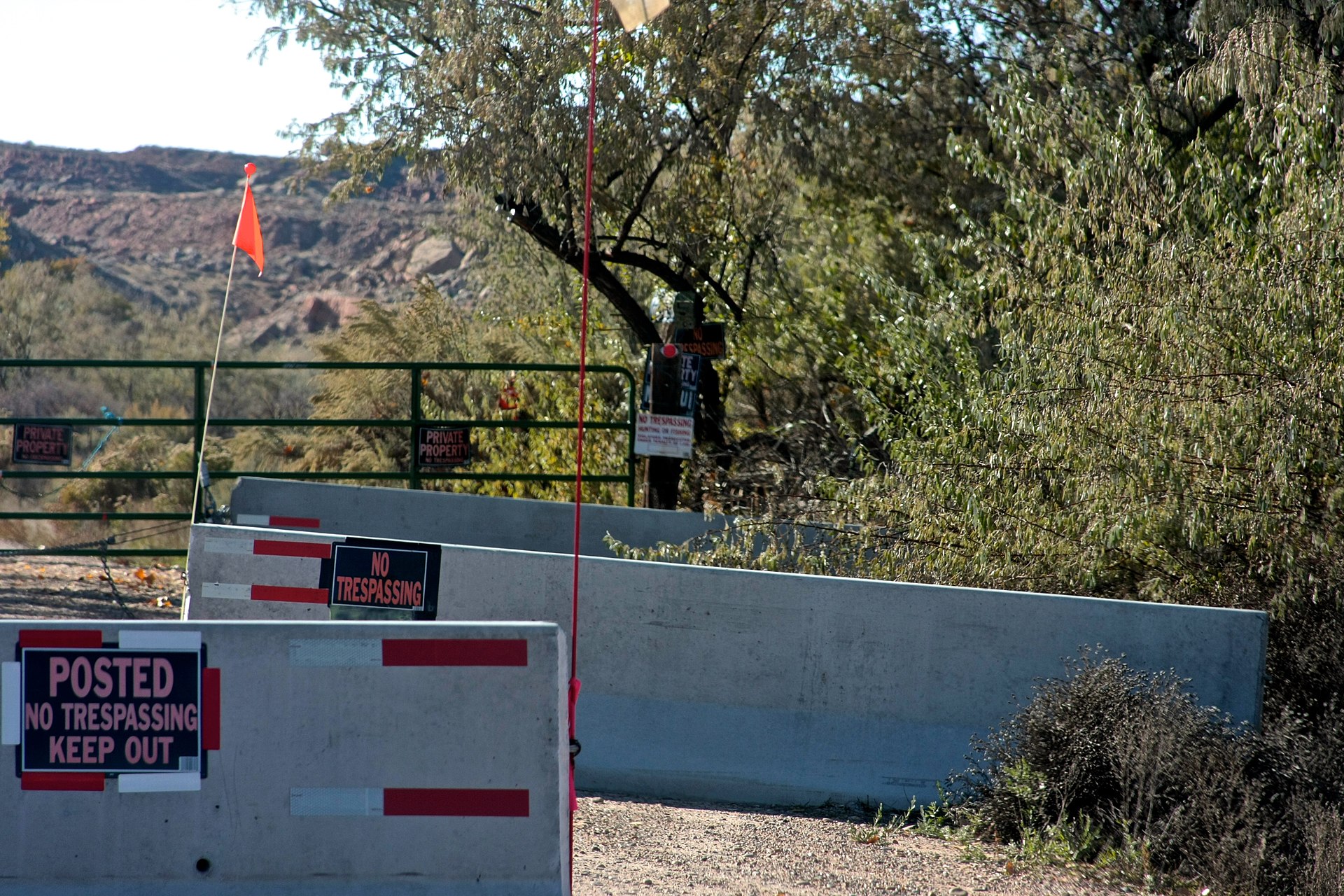 The real front gate of Skinwalker Ranch: POSTED NO TRESPASSING and PRIVATE PROPERTY signs, a concrete barrier and a red flag, with Utah’s red mesa country behind