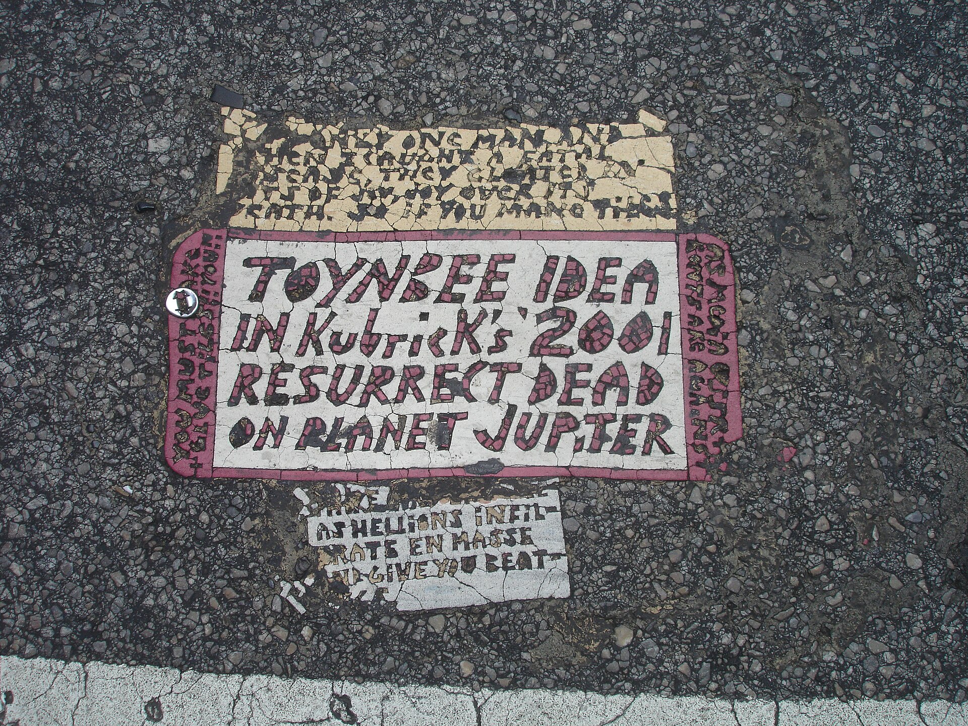 An actual Toynbee tile embedded in the crosswalk at 13th and Market Streets, Philadelphia — with side texts pressed in above and below the main panel