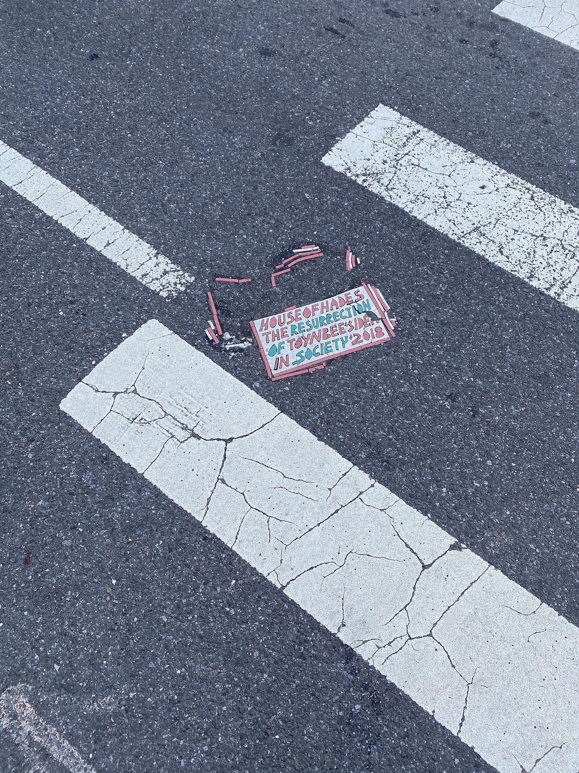 A tile in the crosswalk at W 26th St and 10th Ave, Manhattan — beside the shattered fragments sits a 2018 tile invoking the House of Hades