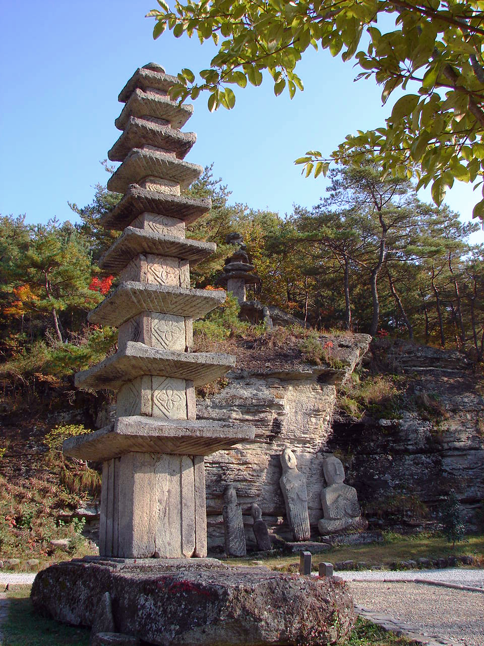 The nine-story stone pagoda at Unjusa in Hwasun, with weathered stone Buddhas lined up beneath the rock cliff behind it — the site as it stands today