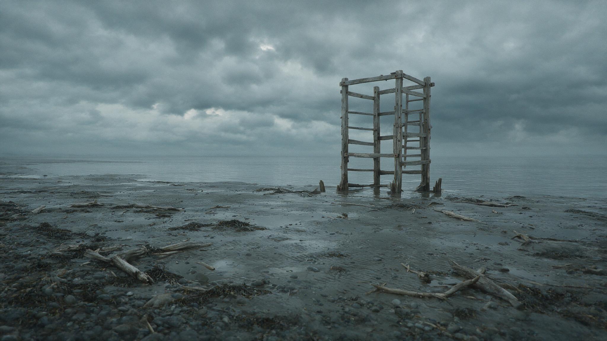 A bleak, overcast shoreline of grey tidal flats and still water under a heavy sky, an abandoned wooden training structure in the distance